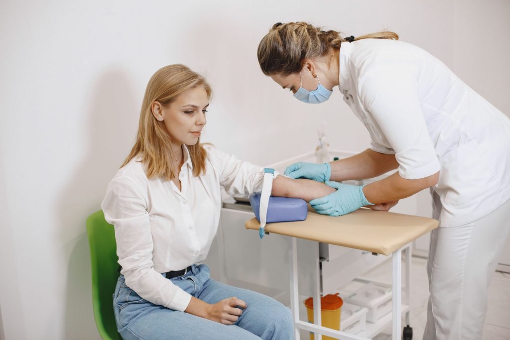 Eine Pflegekraft führt bei einer sitzenden Patientin in einer medizinischen Einrichtung eine Blutabnahme durch.
